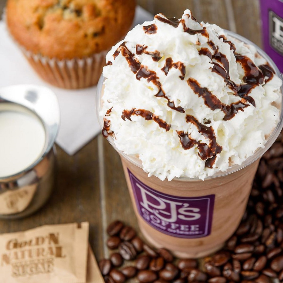 A close up of a PJ's iced specialty drink with whipped cream, a muffin, some coffee beans and a small pitcher of creamer.